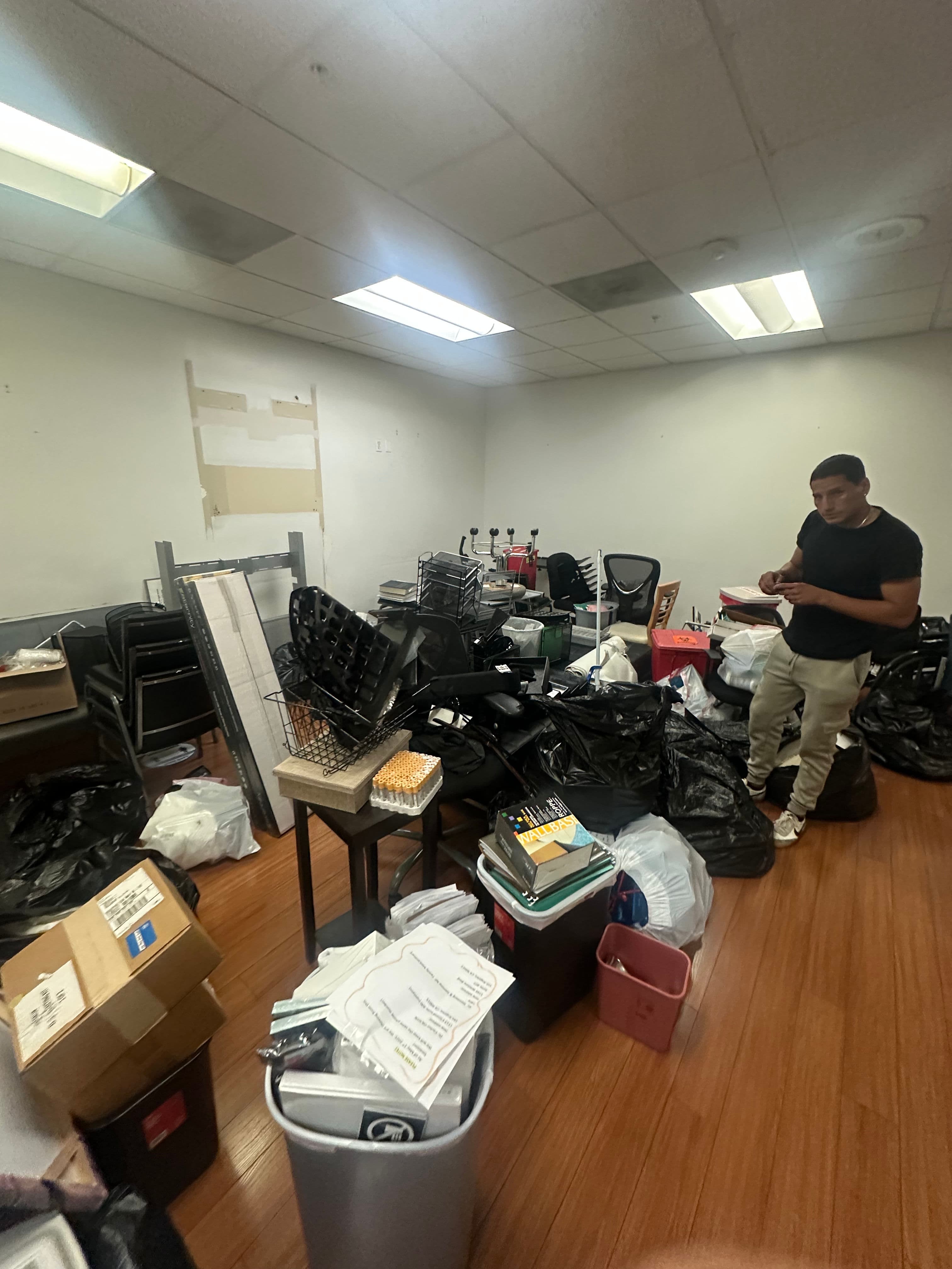 Cluttered office room filled with bags, boxes, and furniture, with a person assessing the space.