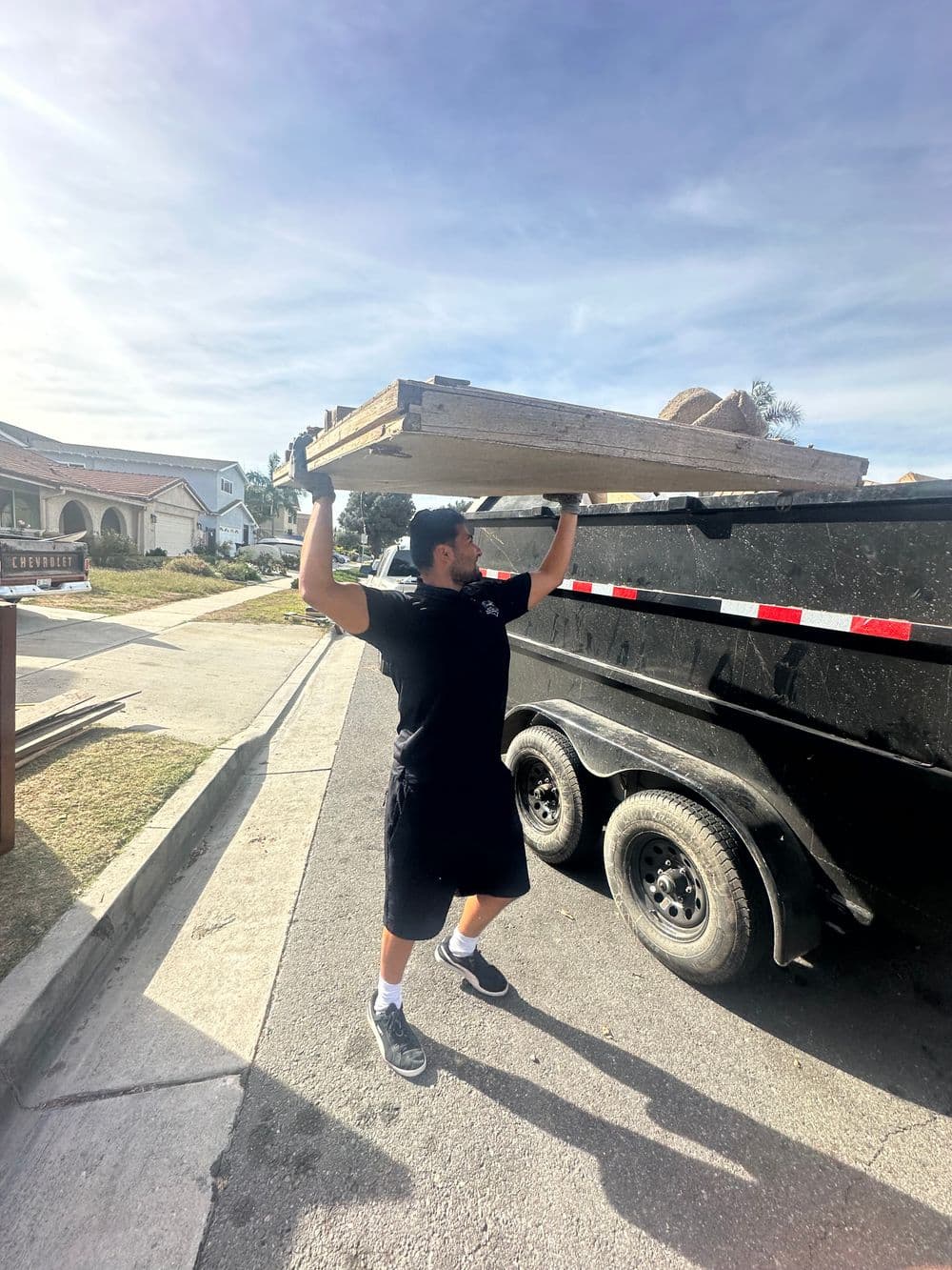 Man lifting a wooden plank above his head while loading a dumpster in a residential area.