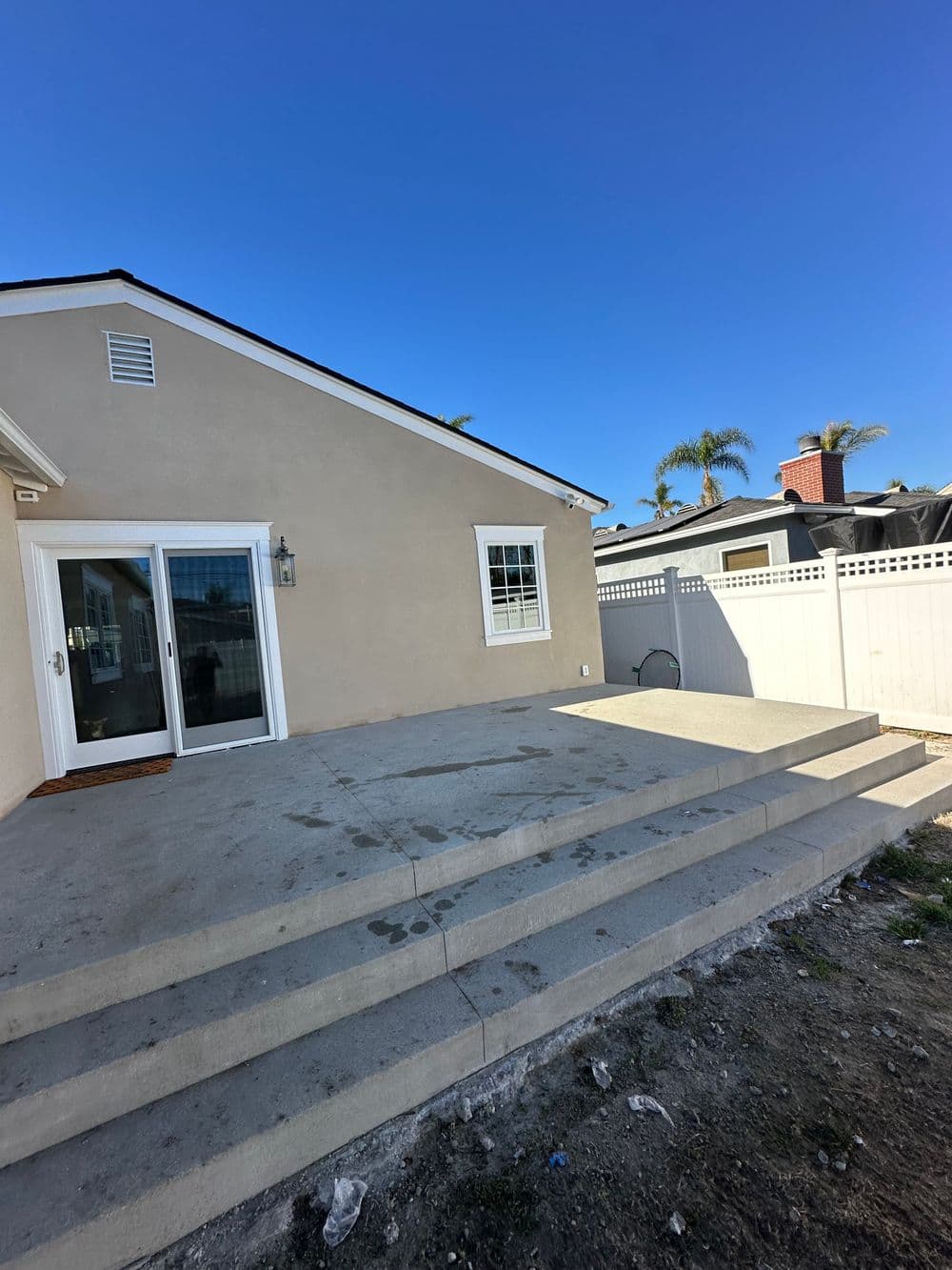 House exterior with steps leading to a patio, surrounded by palm trees and a fence. Clear blue sky.