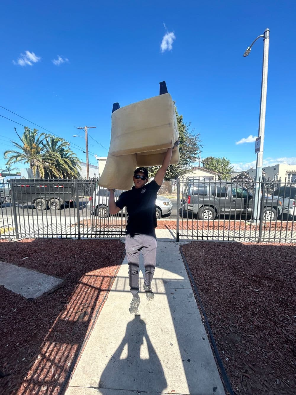 Man carrying large object outdoors with palm trees and vehicles in the background.