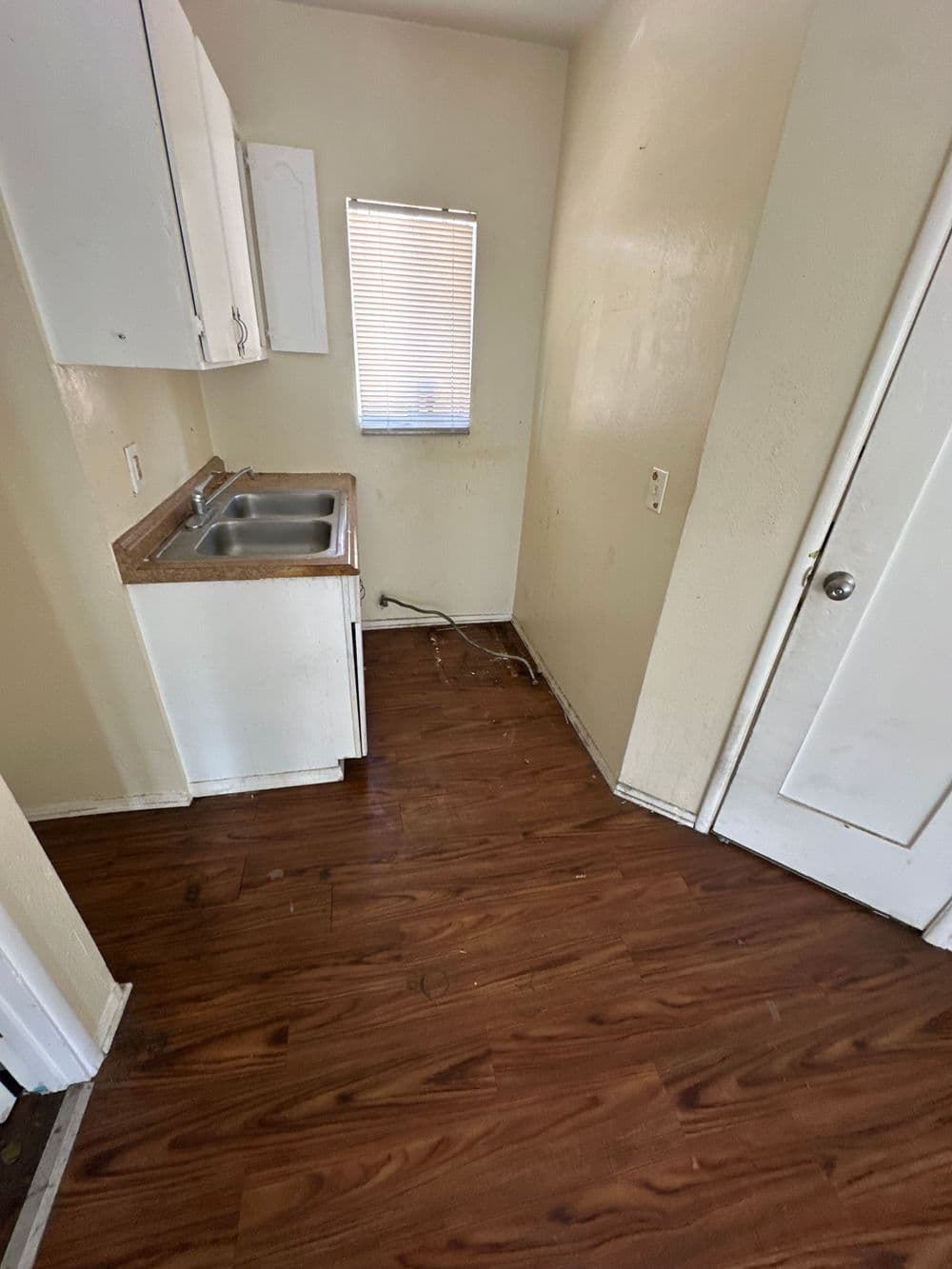 Small kitchen space with wooden flooring, a sink, white cabinets, and a window.