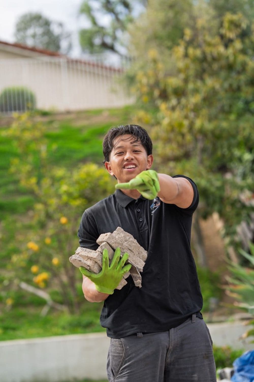 Person in green gloves holding bricks, smiling, outdoors in a garden setting.