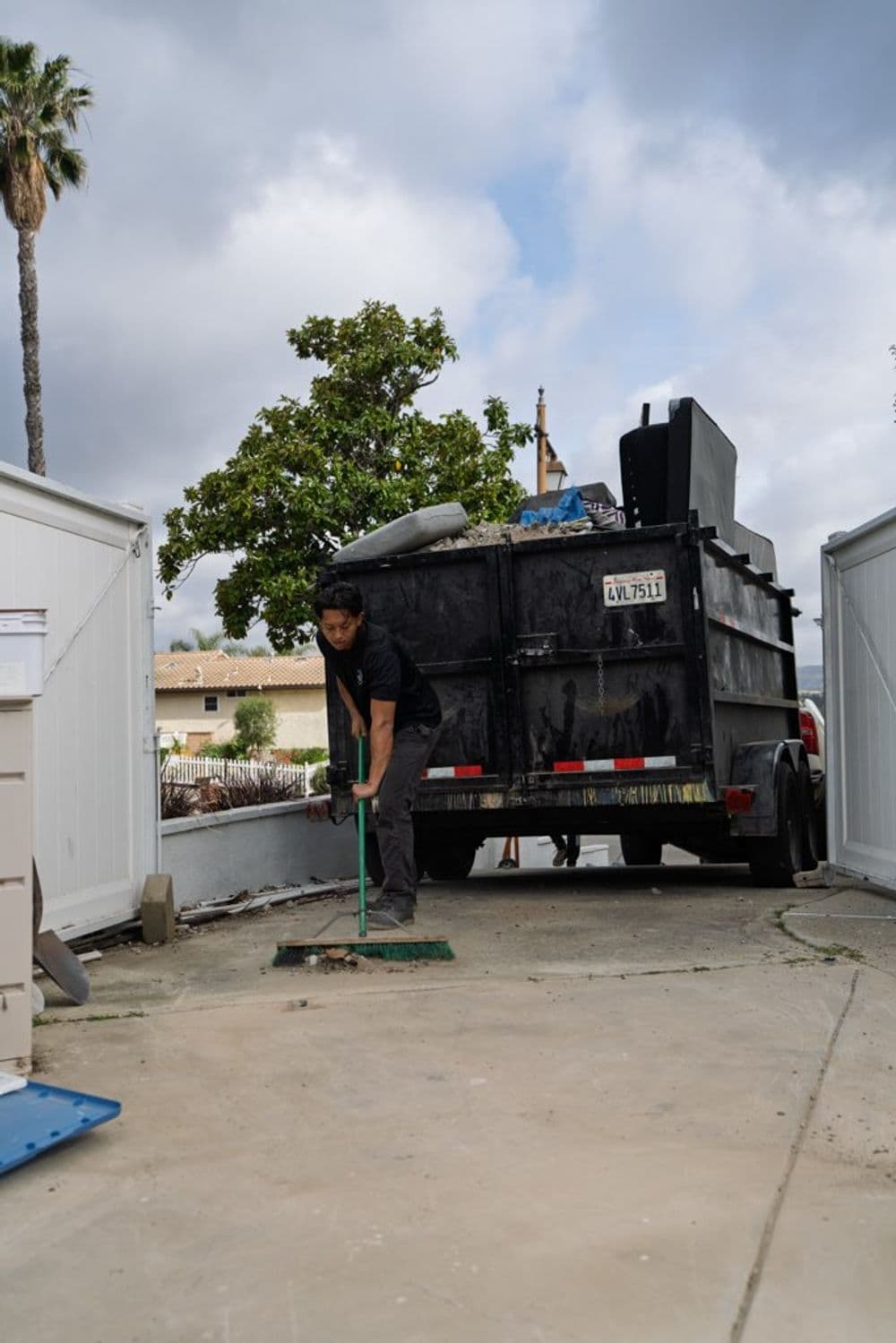 Worker sweeping debris in front of a dumpster on a residential property, cloudy sky.