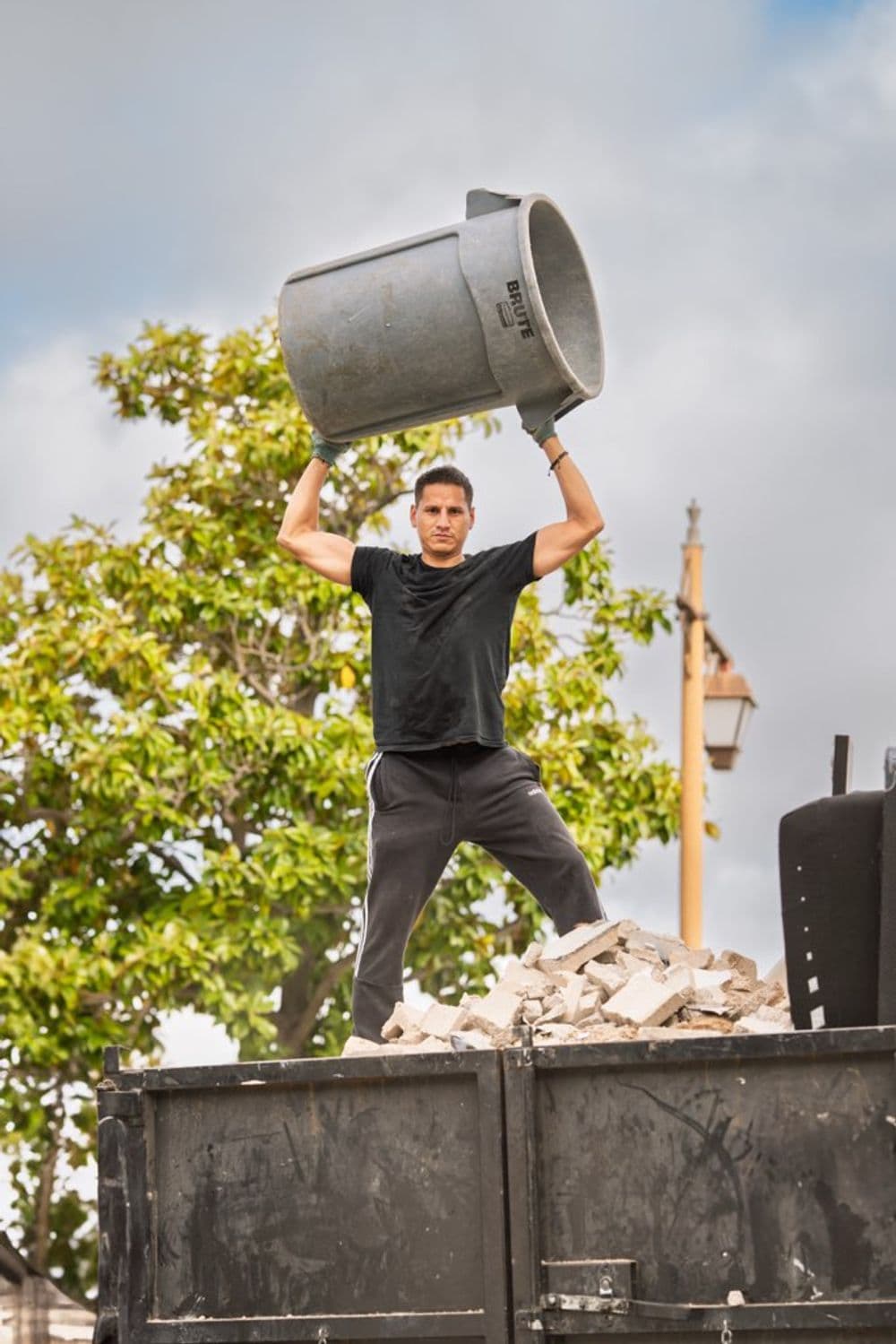 Man lifting a trash bin over his head on a dumpster, trees and lamppost in the background.