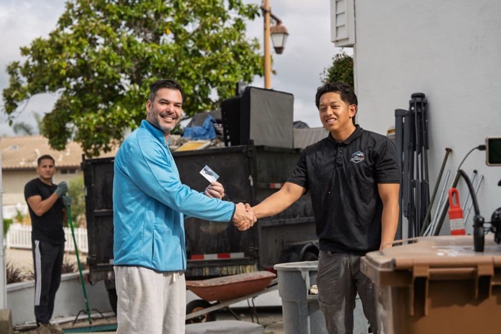 Two men shake hands outside, with a third man working near a dumpster in the background.