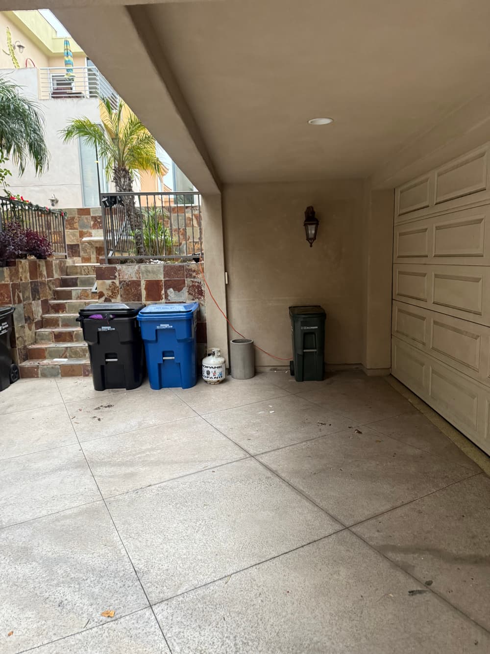 Garbage bins in a residential garage with a stone wall and greenery in the background.