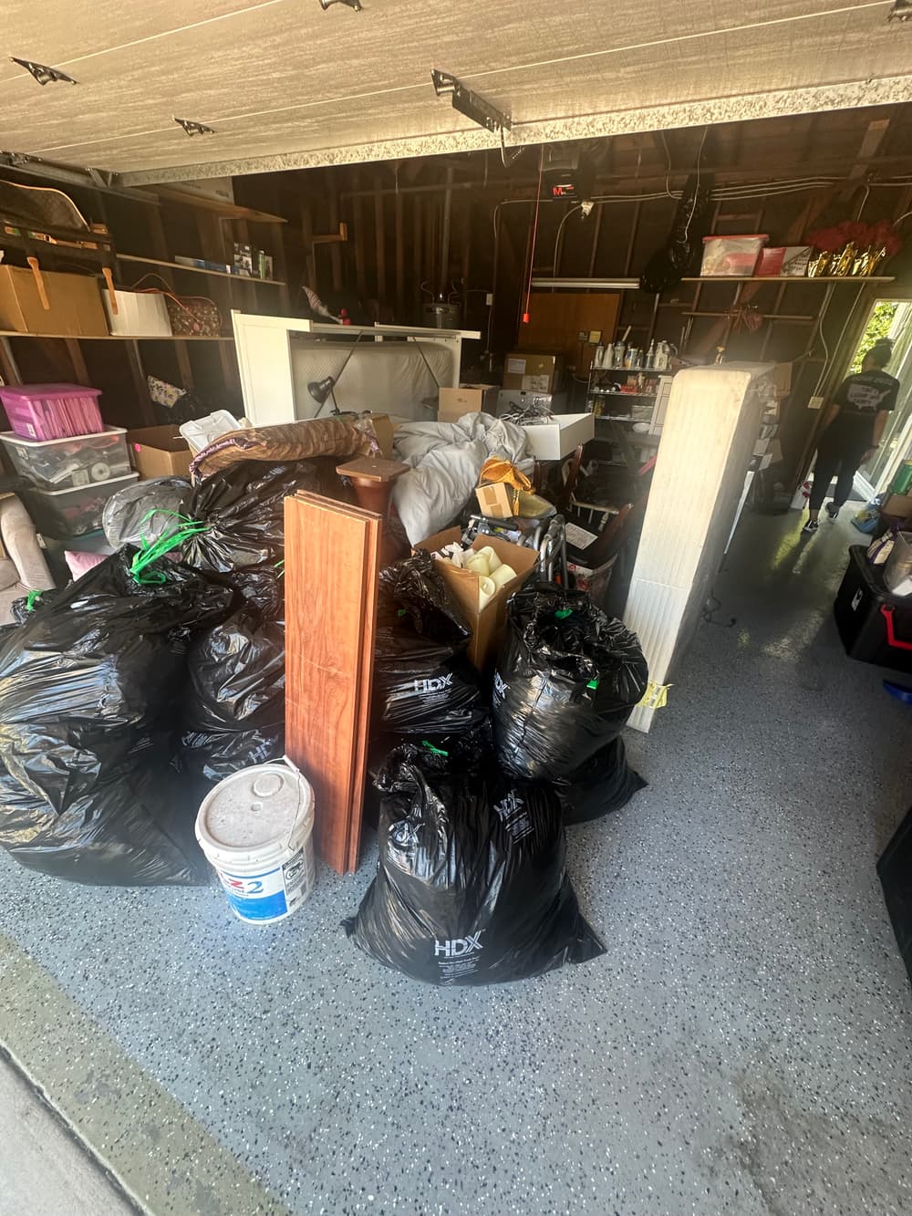 Cluttered garage with black trash bags, storage bins, and household items scattered around.
