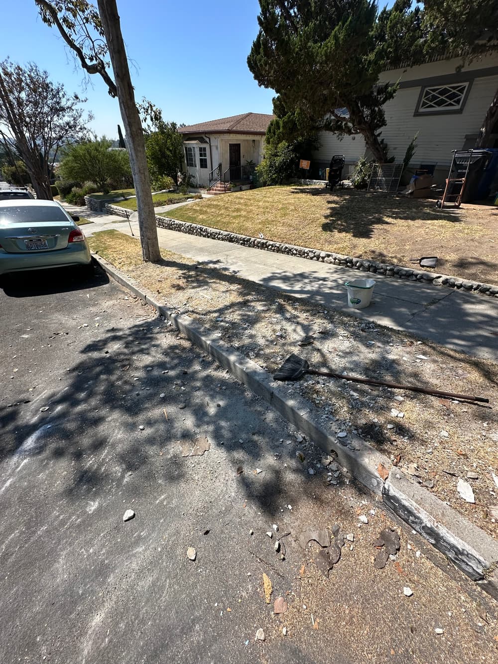 Residential street with a sidewalk under repair, tools scattered, and a house in the background.